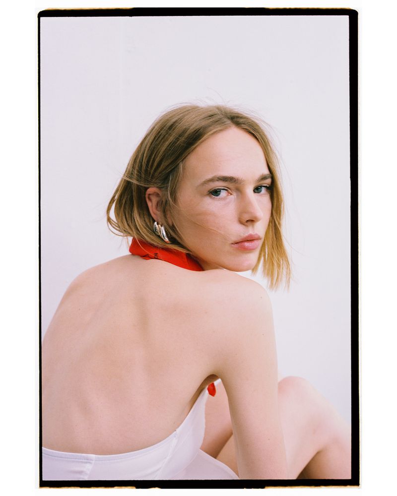 Blonde woman in white dress and red bandana poses in a studio portrait with a blank background.