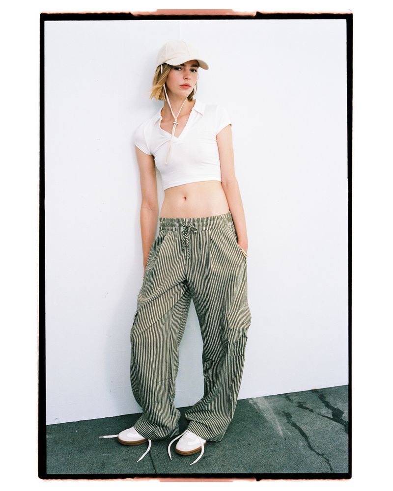 Young woman in casual streetwear stands against a white wall in a full shot, wearing a crop top, striped pants, sneakers, and a baseball cap.