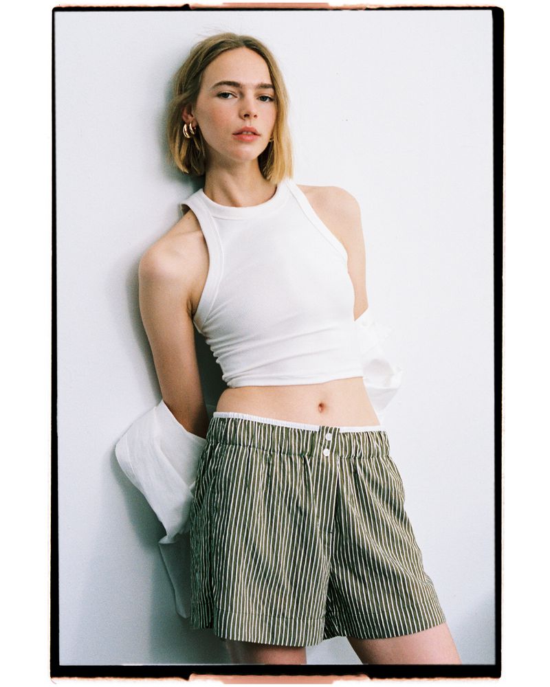 Puck Schrover wearing a white ribbed crop tank top and striped shorts while leaning against a white studio wall.