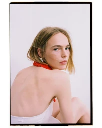 Model Puck Schrover looking over her bare back wearing a red bandana and silver earrings against a white background.