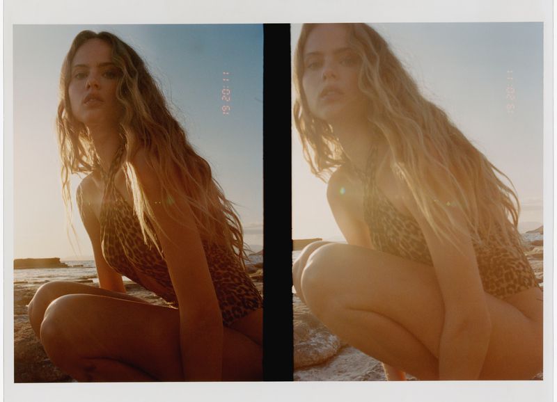 Blonde woman in a leopard print bathing suit crouches on rocks near a beach, bathed in golden light with soft focus and film grain.