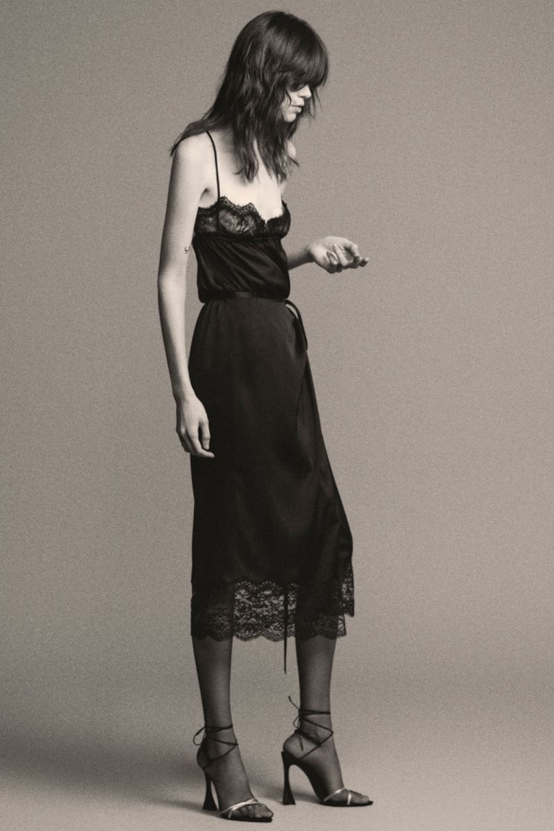 A stylish adult female model poses in a black lace-trimmed slip dress and strappy heels against a neutral backdrop.