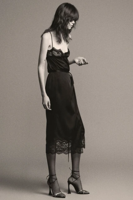 A stylish adult female model poses in a black lace-trimmed slip dress and strappy heels against a neutral backdrop.
