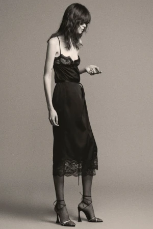 A stylish adult female model poses in a black lace-trimmed slip dress and strappy heels against a neutral backdrop.