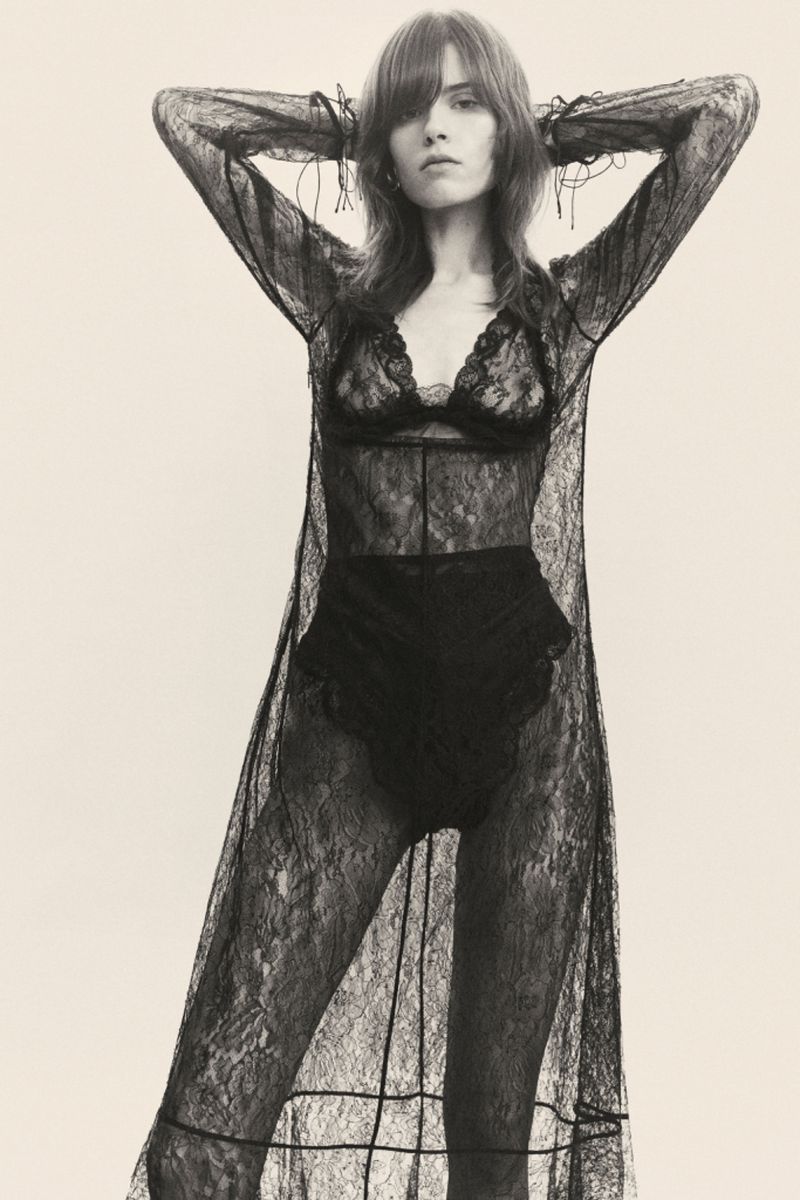 A woman poses in a black lace lingerie set and a long lace robe against a bright background in a studio shot for fashion.