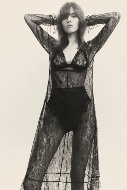 A woman poses in a black lace lingerie set and a long lace robe against a bright background in a studio shot for fashion.