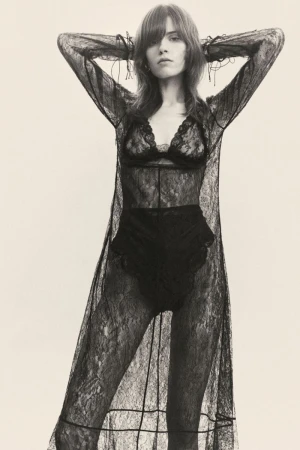 A woman poses in a black lace lingerie set and a long lace robe against a bright background in a studio shot for fashion.
