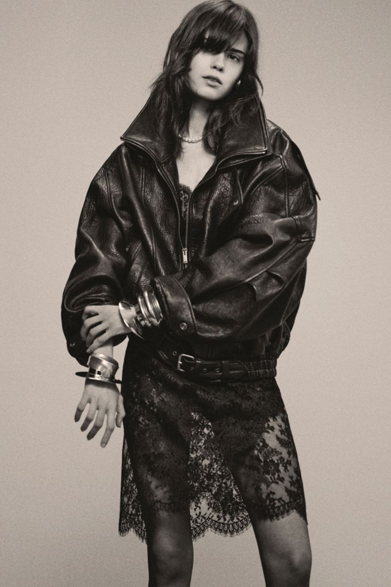 A young adult woman poses in a black leather bomber jacket and lace skirt against a neutral background in a studio fashion shot.