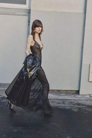 Model walking in a sheer black lace jumpsuit and stiletto heels holding a glossy black coat outdoors against a grey wall.