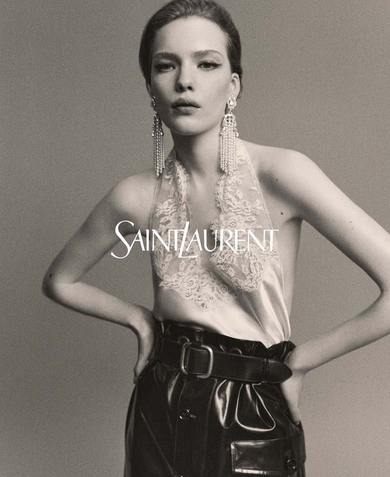 Woman poses in a studio wearing dangling earrings, a lace top, and high-waisted leather trousers for a Saint Laurent advertisement.