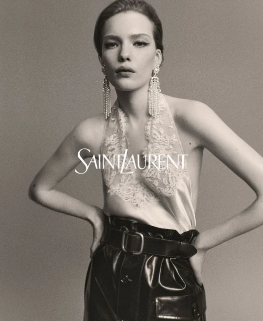 Woman poses in a studio wearing dangling earrings, a lace top, and high-waisted leather trousers for a Saint Laurent advertisement.