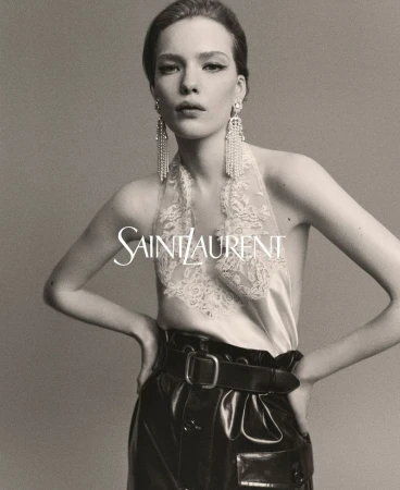 Woman poses in a studio wearing dangling earrings, a lace top, and high-waisted leather trousers for a Saint Laurent advertisement.