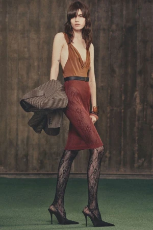 Fashion model poses in brown top, patterned skirt and hosiery, and heels with a grey jacket indoors against a textured background.