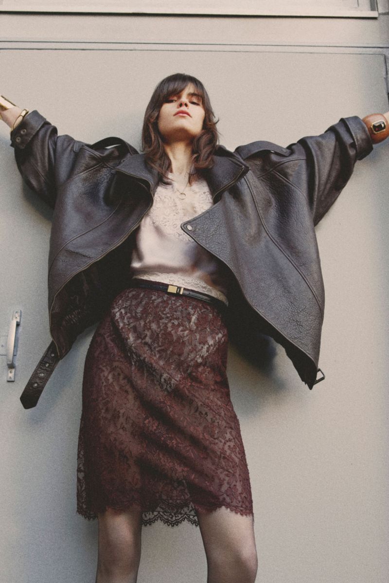 A woman poses in an oversized leather jacket and burgundy lace skirt against a neutral wall, creating an editorial fashion image.