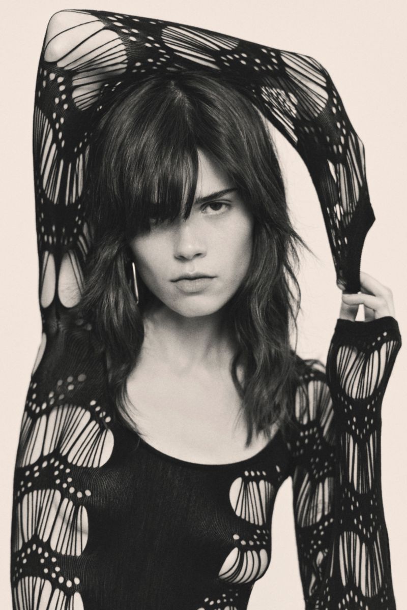 Portrait of a woman with long brown hair wearing a black, long-sleeved mesh top against a cream background in a studio setting.