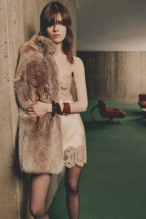 Model leaning against a wall wearing a beige lace slip dress, sheer tights, and a plush fur coat over one shoulder.