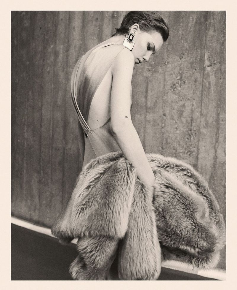 Woman in low-back dress holds fur coat against concrete wall in muted light, accessorized with square earrings, fashion editorial.