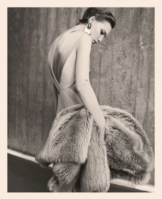 Woman in low-back dress holds fur coat against concrete wall in muted light, accessorized with square earrings, fashion editorial.