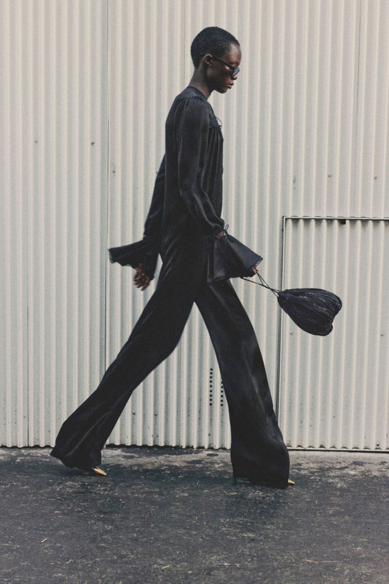 An adult fashion model walks in a black outfit, wide-legged pants, and a bag against a light-colored industrial background, outdoors.