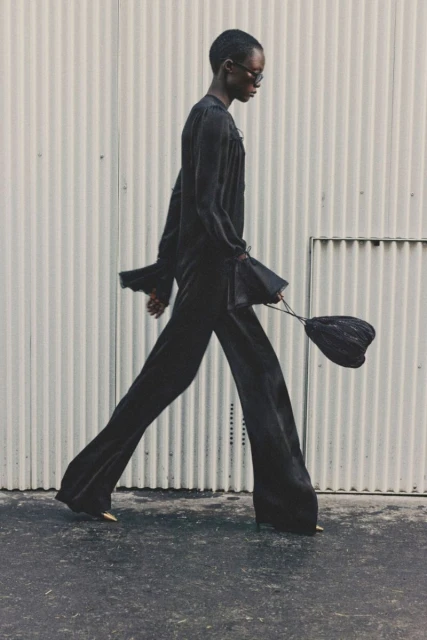 An adult fashion model walks in a black outfit, wide-legged pants, and a bag against a light-colored industrial background, outdoors.