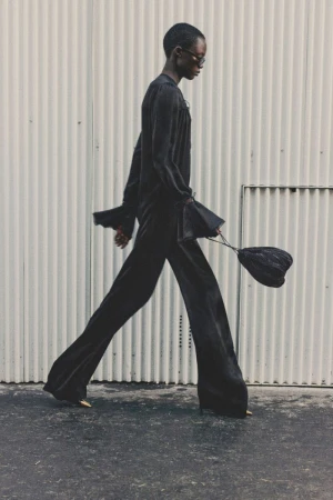 An adult fashion model walks in a black outfit, wide-legged pants, and a bag against a light-colored industrial background, outdoors.