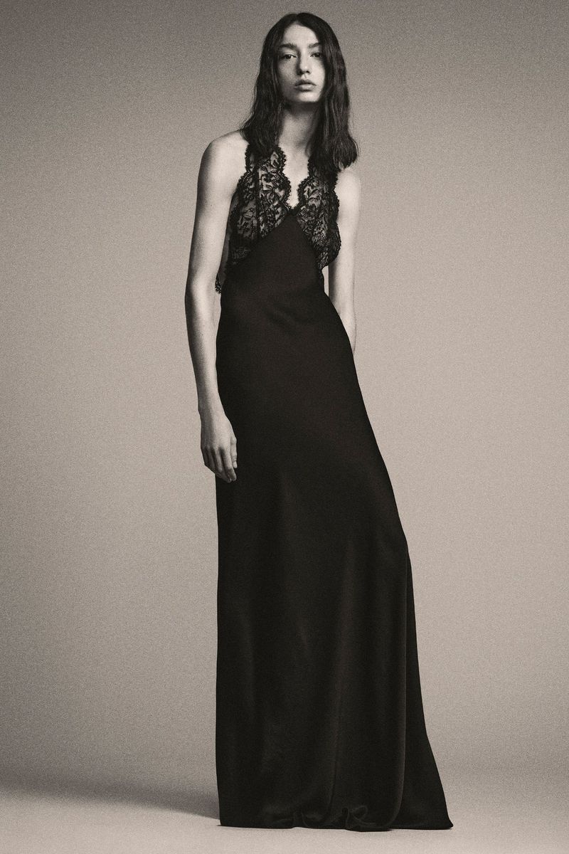 A woman poses in a full-length black gown with lace detail on a light grey background, showcasing fashion and style in a studio setting.