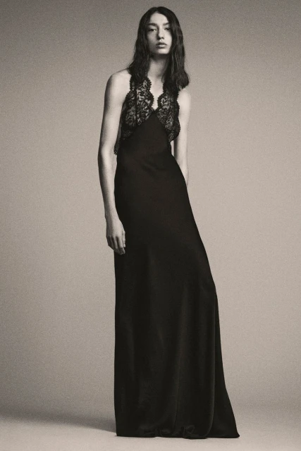A woman poses in a full-length black gown with lace detail on a light grey background, showcasing fashion and style in a studio setting.