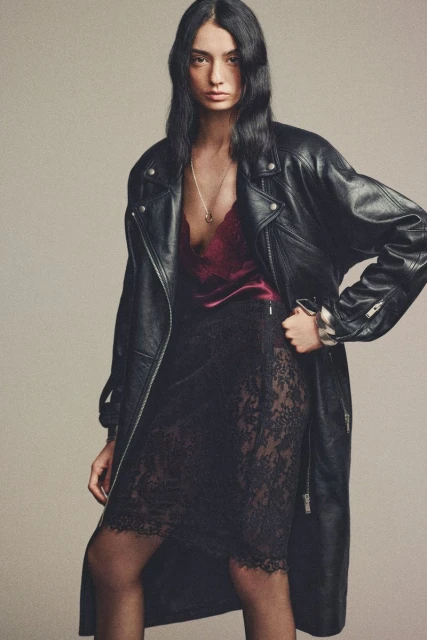 A fashionable woman with long hair wearing a long black leather jacket, dark red silk top, and black lace skirt stands in a studio.