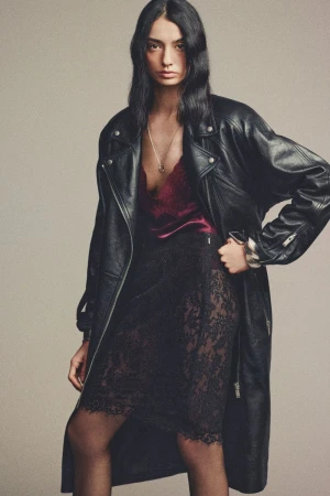 A fashionable woman with long hair wearing a long black leather jacket, dark red silk top, and black lace skirt stands in a studio.