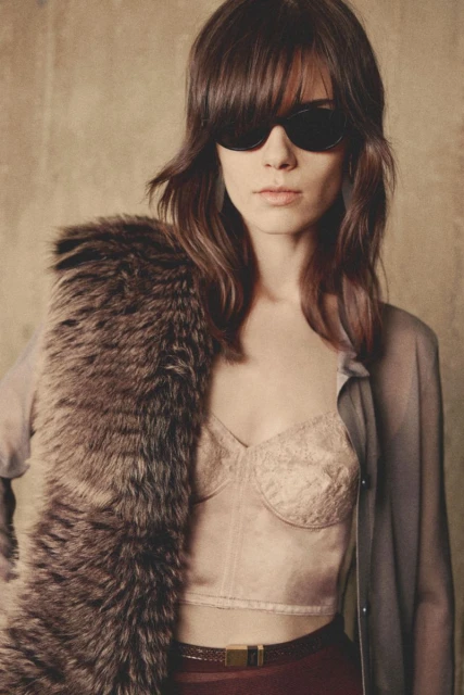 Portrait of a woman with dark sunglasses and a fur stole. She is wearing a lacy top and a maroon skirt with a thin belt.