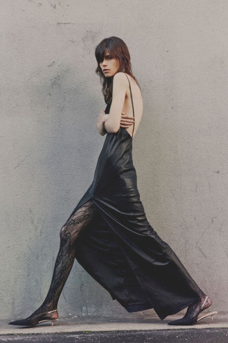 A slender woman in a black dress and patterned tights walks against a neutral wall in a fashion editorial shot with natural light.