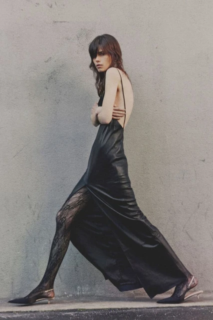 A slender woman in a black dress and patterned tights walks against a neutral wall in a fashion editorial shot with natural light.