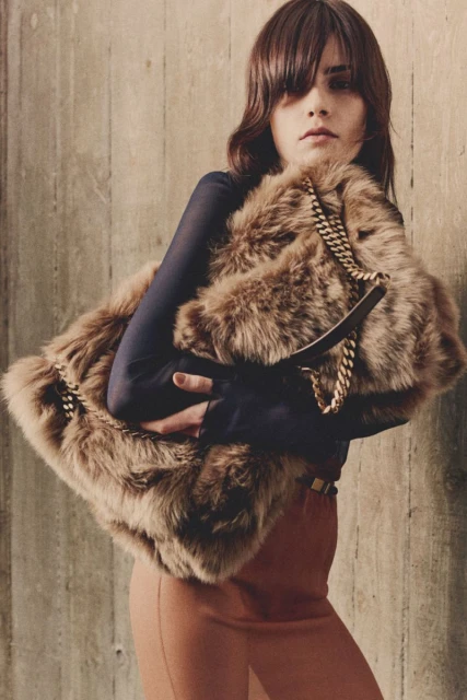 A stylish adult female poses indoors with a large brown faux-fur handbag and a long-sleeve top, complemented by brown trousers and a gold belt.