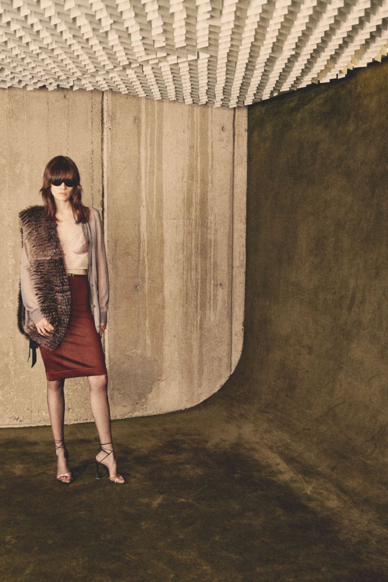 Indoor fashion shot featuring an adult female model wearing sunglasses, a fur stole, a skirt, and heels, posing against a concrete wall.
