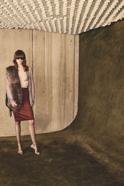 Indoor fashion shot featuring an adult female model wearing sunglasses, a fur stole, a skirt, and heels, posing against a concrete wall.