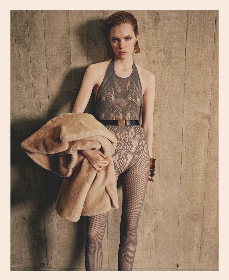 A woman in a floral halter bodysuit and fishnet tights poses against a wooden wall, holding a tan coat. Fashion editorial shot.