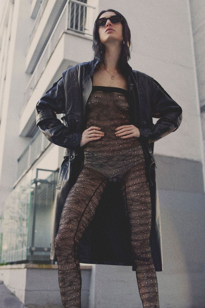 A woman in a sheer bodysuit and leather coat stands confidently against an urban backdrop, looking into the distance with sunglasses.
