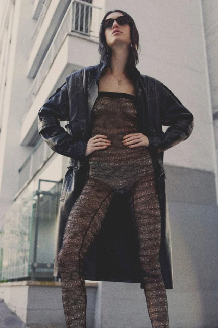 A woman in a sheer bodysuit and leather coat stands confidently against an urban backdrop, looking into the distance with sunglasses.
