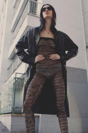 A woman in a sheer bodysuit and leather coat stands confidently against an urban backdrop, looking into the distance with sunglasses.
