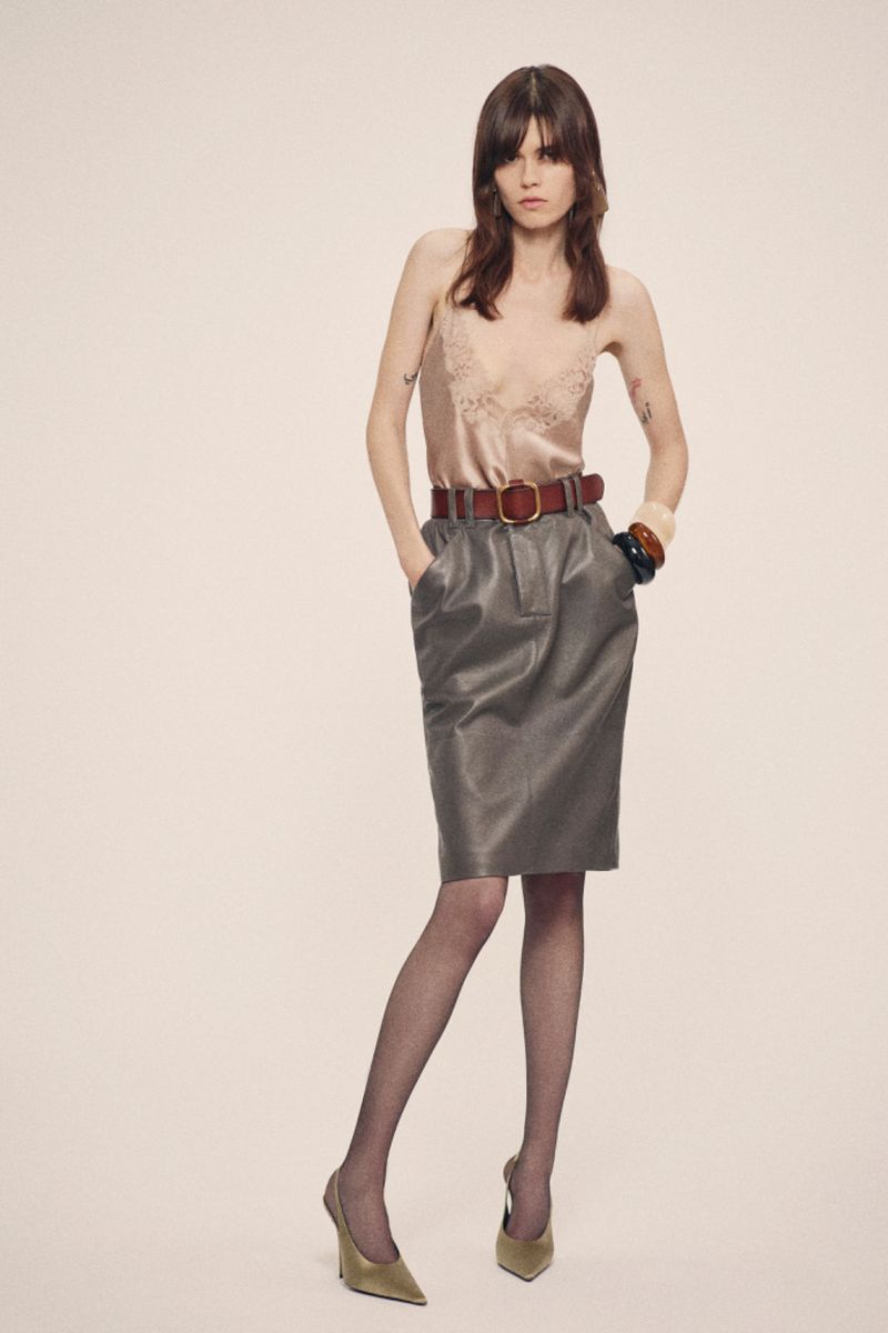 A woman poses in a satin top, leather skirt, sheer tights, and heels in a studio, showcasing a blend of classic and modern fashion.