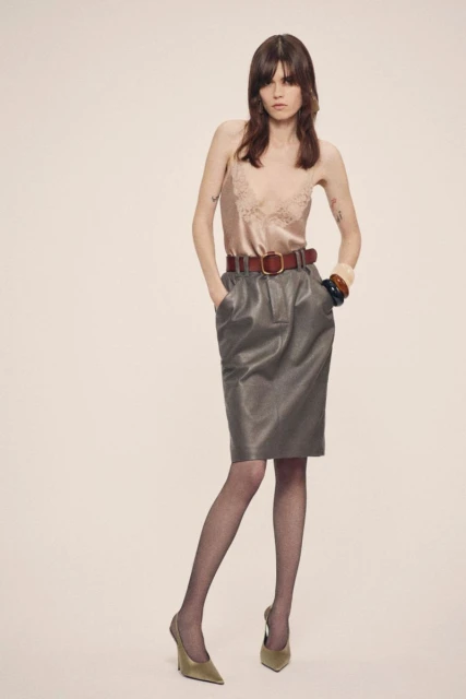 A woman poses in a satin top, leather skirt, sheer tights, and heels in a studio, showcasing a blend of classic and modern fashion.