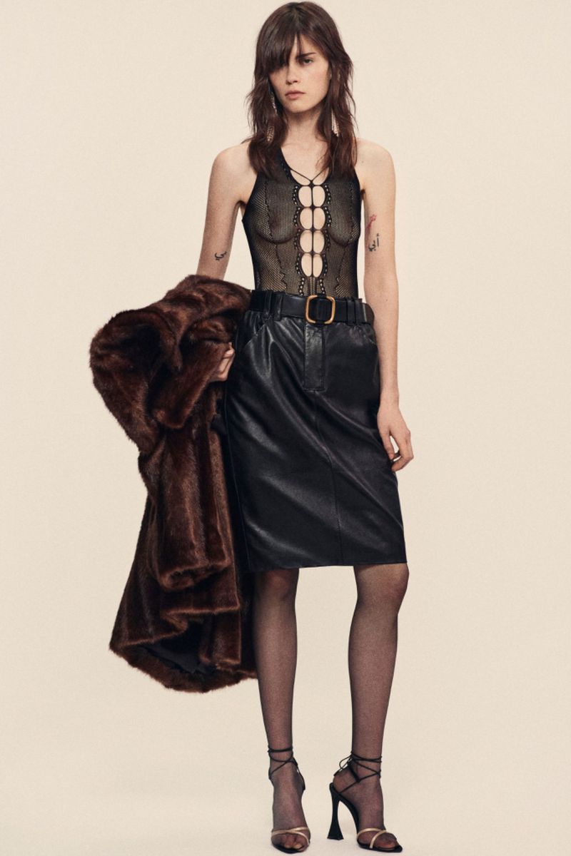 A woman poses in a black lace halter top and leather skirt, holding a brown fur coat against a beige background in a fashion shot.