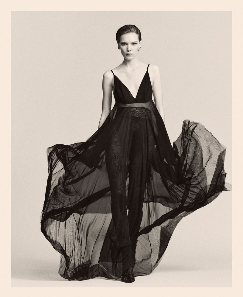 Elegant woman in flowing black dress posing in studio. Fashion shot featuring sheer fabric, black and white tones, and subtle lighting.