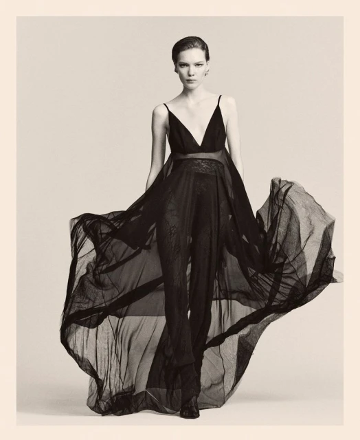 Elegant woman in flowing black dress posing in studio. Fashion shot featuring sheer fabric, black and white tones, and subtle lighting.