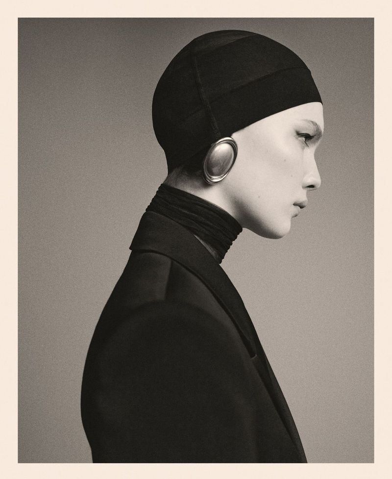 Striking portrait of a woman in a black head covering and jacket, accented by oval earrings, against a neutral backdrop. Fashion-forward.
