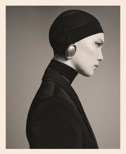 Striking portrait of a woman in a black head covering and jacket, accented by oval earrings, against a neutral backdrop. Fashion-forward.