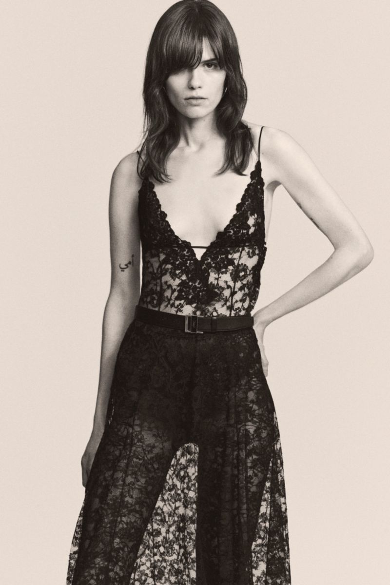 A woman poses in a black lace bodysuit and pants with a belt, styled with long brunette hair and bangs against a neutral backdrop.