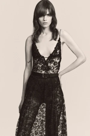 A woman poses in a black lace bodysuit and pants with a belt, styled with long brunette hair and bangs against a neutral backdrop.
