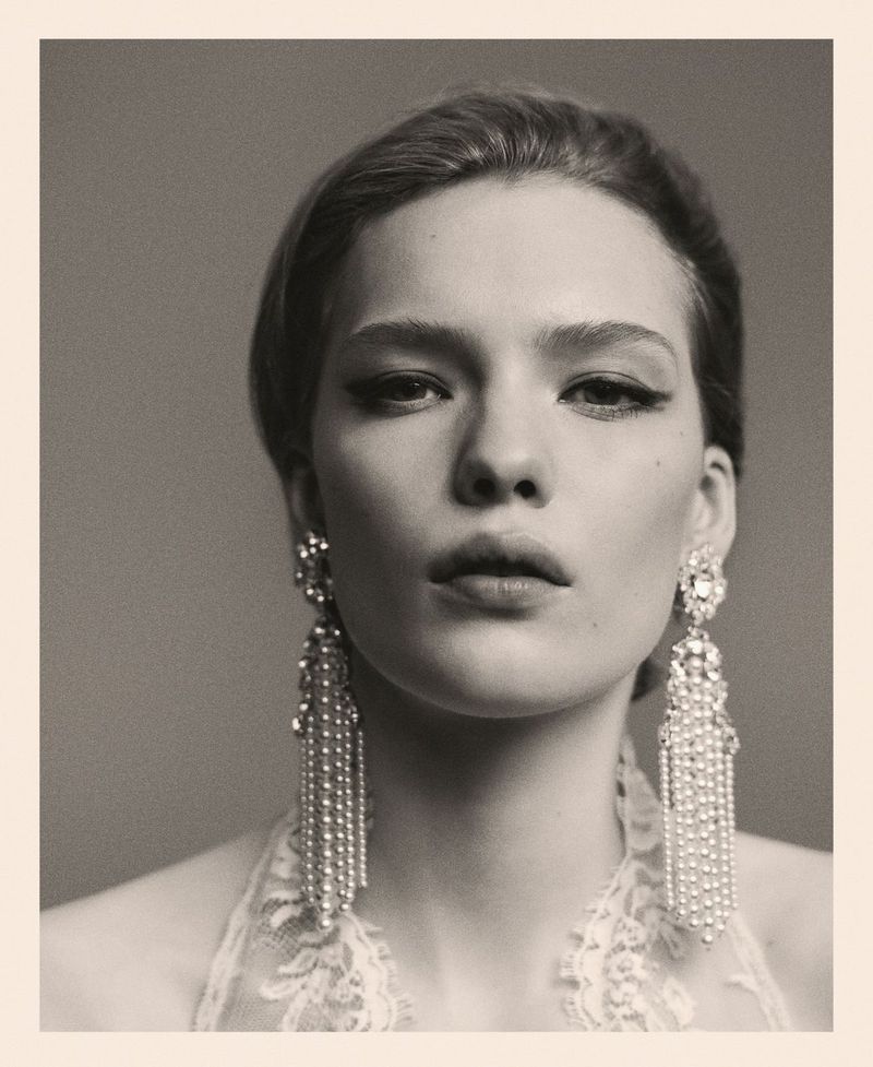 Close-up portrait of a stylish woman wearing crystal earrings and elegant makeup, set against a grey background in muted tones.