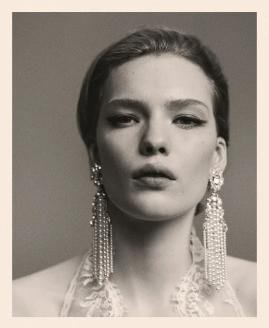 Close-up portrait of a stylish woman wearing crystal earrings and elegant makeup, set against a grey background in muted tones.